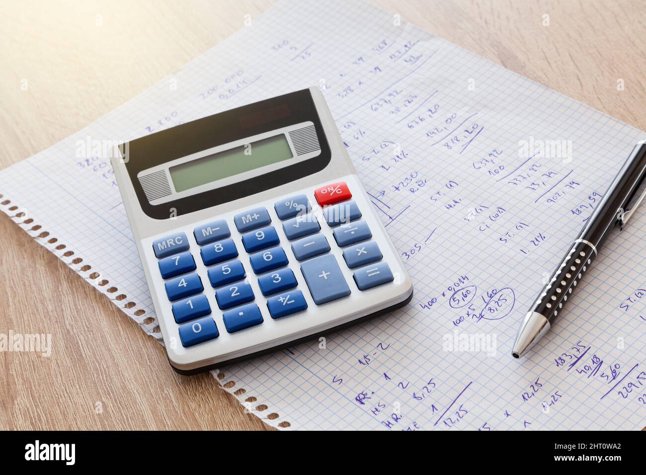 Sur une table en bois se trouve une feuille de carrés de bloc-notes avec des nombres et des calculs manuscrits et sur la feuille se trouve un stylo et une calculatrice. Banque D'Images