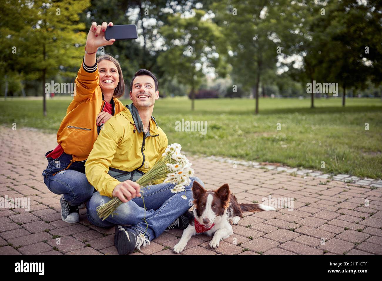 belle jeune femme en tenue décontractée prenant le selfie d'elle-même et un petit ami assis avec les jambes croisées et un chien au milieu d'un chemin dans le parc Banque D'Images