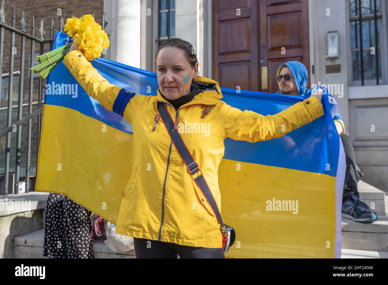 Londres, Royaume-Uni. 26 février 2022. Une femme se tient debout devant un grand drapeau ukrainien lors d'une manifestation publique au Royaume-Uni, tenant un bouquet de fleurs jaunes et portant une veste jaune avec des accents bleus. Sa joue est peinte d'une bande bleue et jaune, symbolisant le soutien à l'Ukraine. Une autre participante est visible derrière elle, elle aussi vêtue de couleurs ukrainiennes. La scène capture un moment de solidarité, de fierté nationale et d’expression civique pacifique. Penelope Barritt/Alamy Live News Banque D'Images