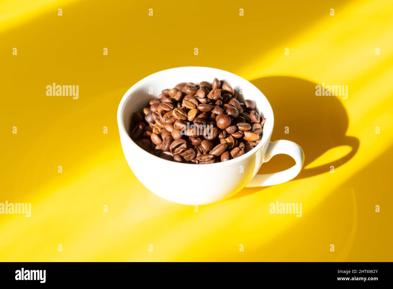Grains de café dans une tasse en céramique blanche sur fond jaune. Ombres dures du soleil Banque D'Images