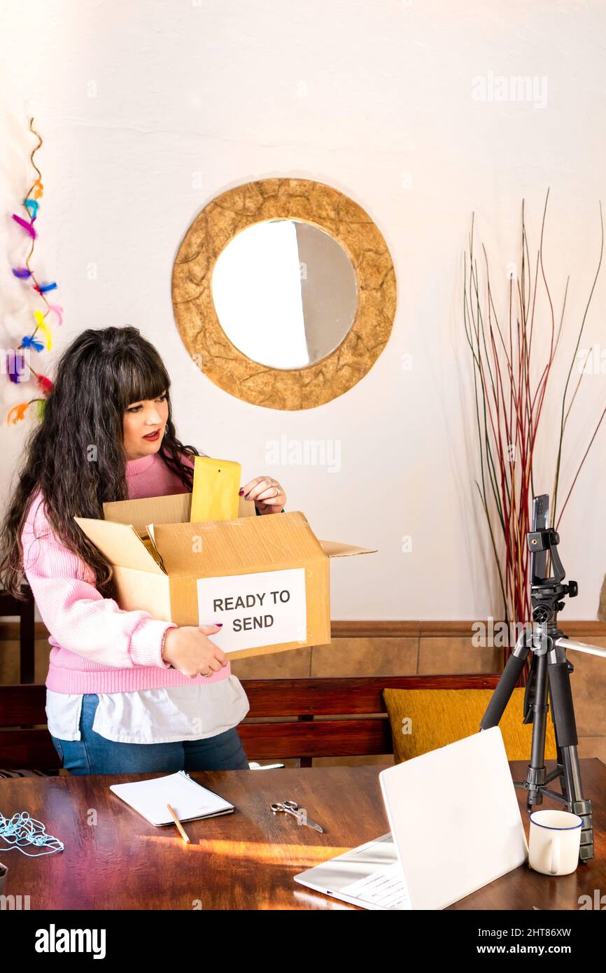 Femme blogger emballage enveloppes avec commandes dans carton pour livraison tout en enregistrant la vidéo sur téléphone portable attaché sur trépied dans l'atelier d'artisanat Banque D'Images