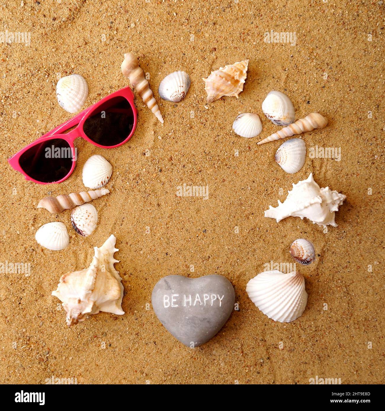 Une pierre en forme de coeur avec l'inscription être heureux, des lunettes et divers types de coquillages se trouvent sur le sable de plage en forme de cercle. Dans Banque D'Images