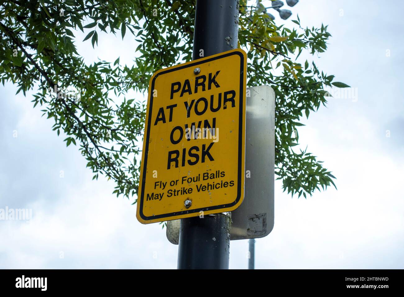 Vue à angle bas d'un parc à votre propre panneau de risque à l'extérieur d'un terrain de baseball, avertissant quiconque se garer sa voiture de la possibilité de balles de mouche Banque D'Images