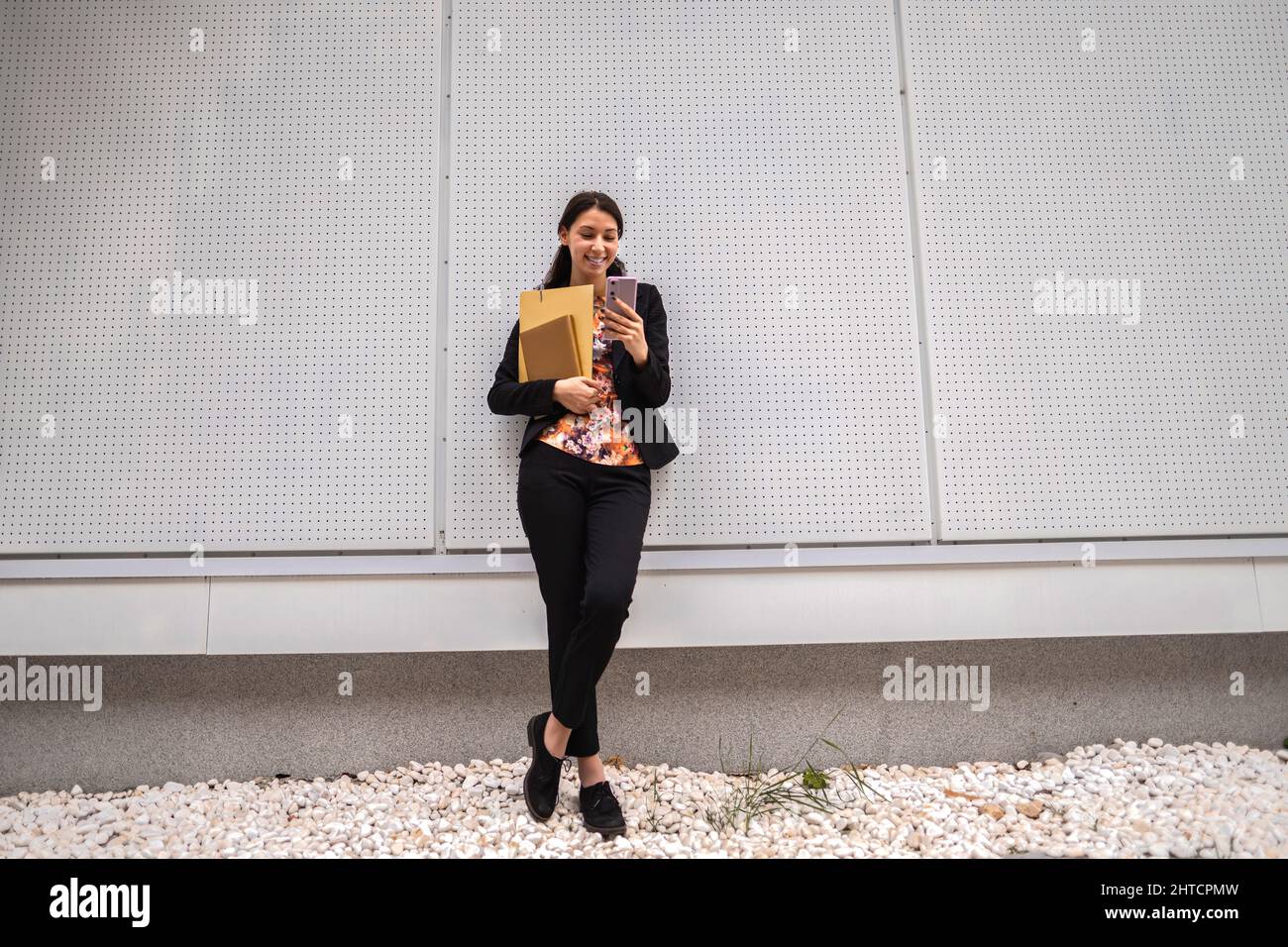 Jeune femme caucasienne joyeuse dans une tenue chic et décontractée, penchée sur un mur à l'aide de son smartphone Banque D'Images