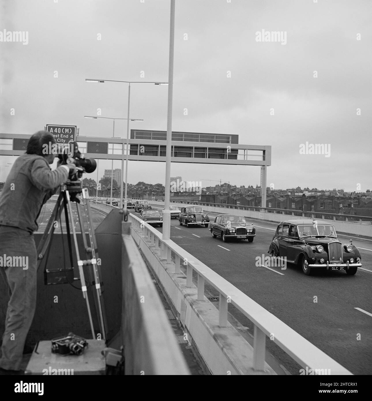 Westway Flyover, A40, Paddington, Cité de Westminster, Londres, 28/07/1970. Un homme filmant un convoi de voitures officielles à l'ouverture du survol de Westway depuis l'arrière d'un camion sur la chaussée opposée. Les travaux sur le site de l'extension de l'avenue Western ont commencé le 1st septembre 1966 et le Westway, tel qu'il a été connu, a été officiellement ouvert le 28th juillet 1970. L'autoroute en hauteur reliant la A40 à White City à Marylebone Road à Paddington, à environ 2 et#xbd; Miles, était la plus longue d'Europe. Les ingénieurs-conseils G Maunsell & amp ; les partenaires ont conçu le projet routier pour le Grand London Counc Banque D'Images