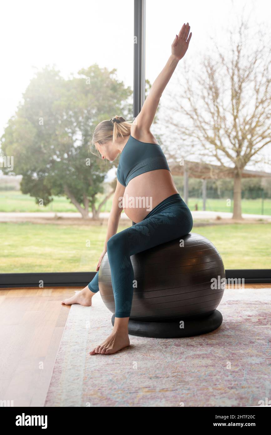 Femme enceinte blonde pratiquant le yoga dans le salon à côté des fenêtres Banque D'Images