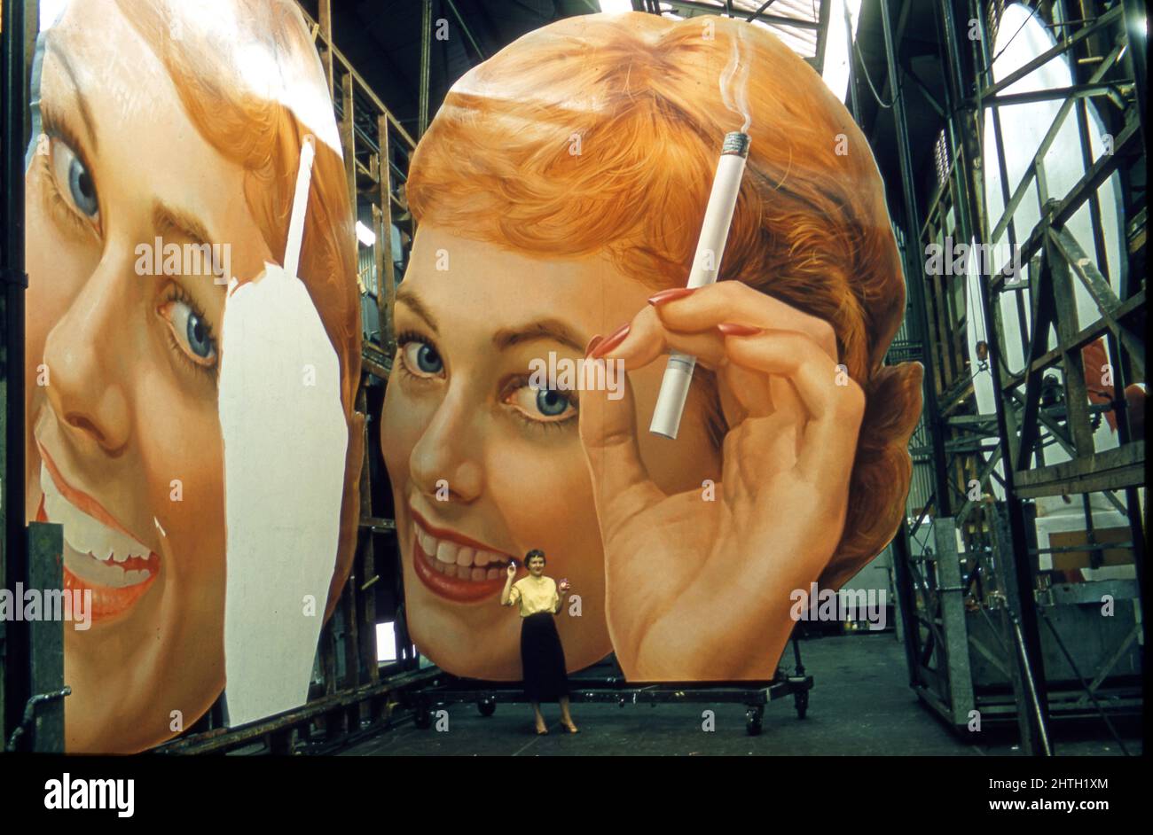 Une femme qui fume une cigarette est éclipsé par la peinture de panneaux publicitaires dans un studio de publicité en plein air à Londres, en Angleterre vers 1950s. Banque D'Images