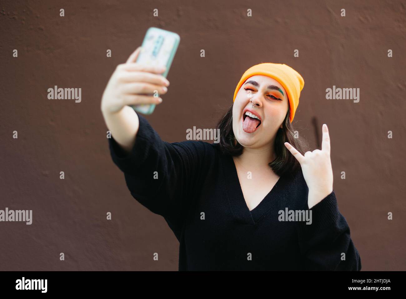 Jeune femme montrant le mouvement du rock et du roulis et montrant la langue tout en prenant autoportrait sur smartphone sur fond marron en ville Banque D'Images