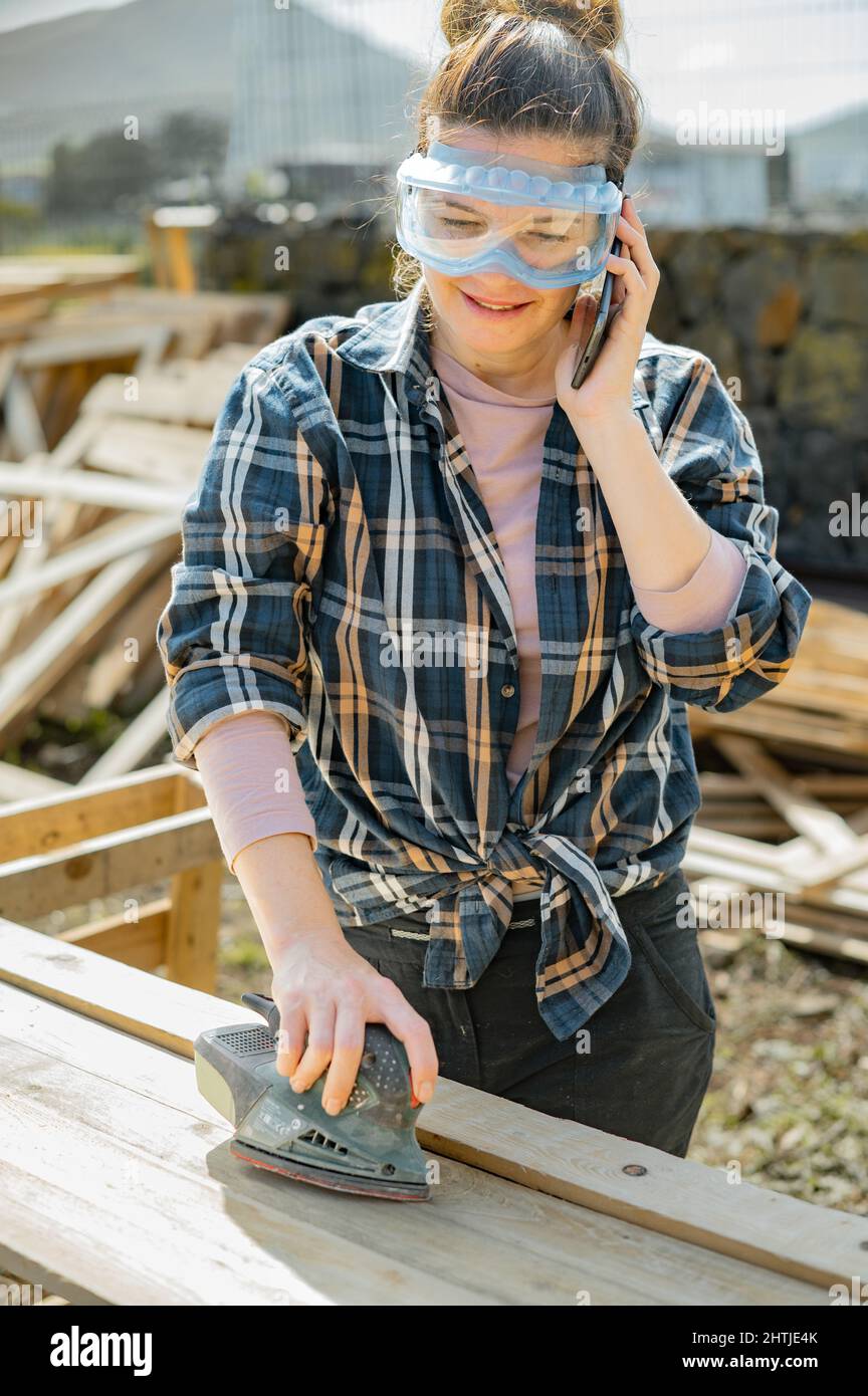 Femme en lunettes de protection téléphone parlant tout en ponçant des planches en bois avec une ponceuse orbitale dans la campagne le jour d'été Banque D'Images