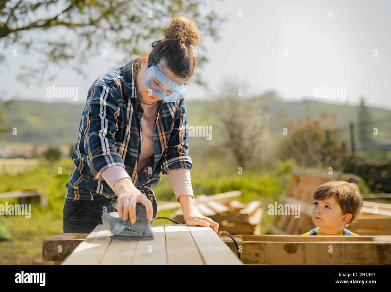 Femme concentrée dans des verres de protection ponçant des planches en bois avec une ponceuse électrique spéciale tout en se tenant près de mignon garçon dans la campagne Banque D'Images