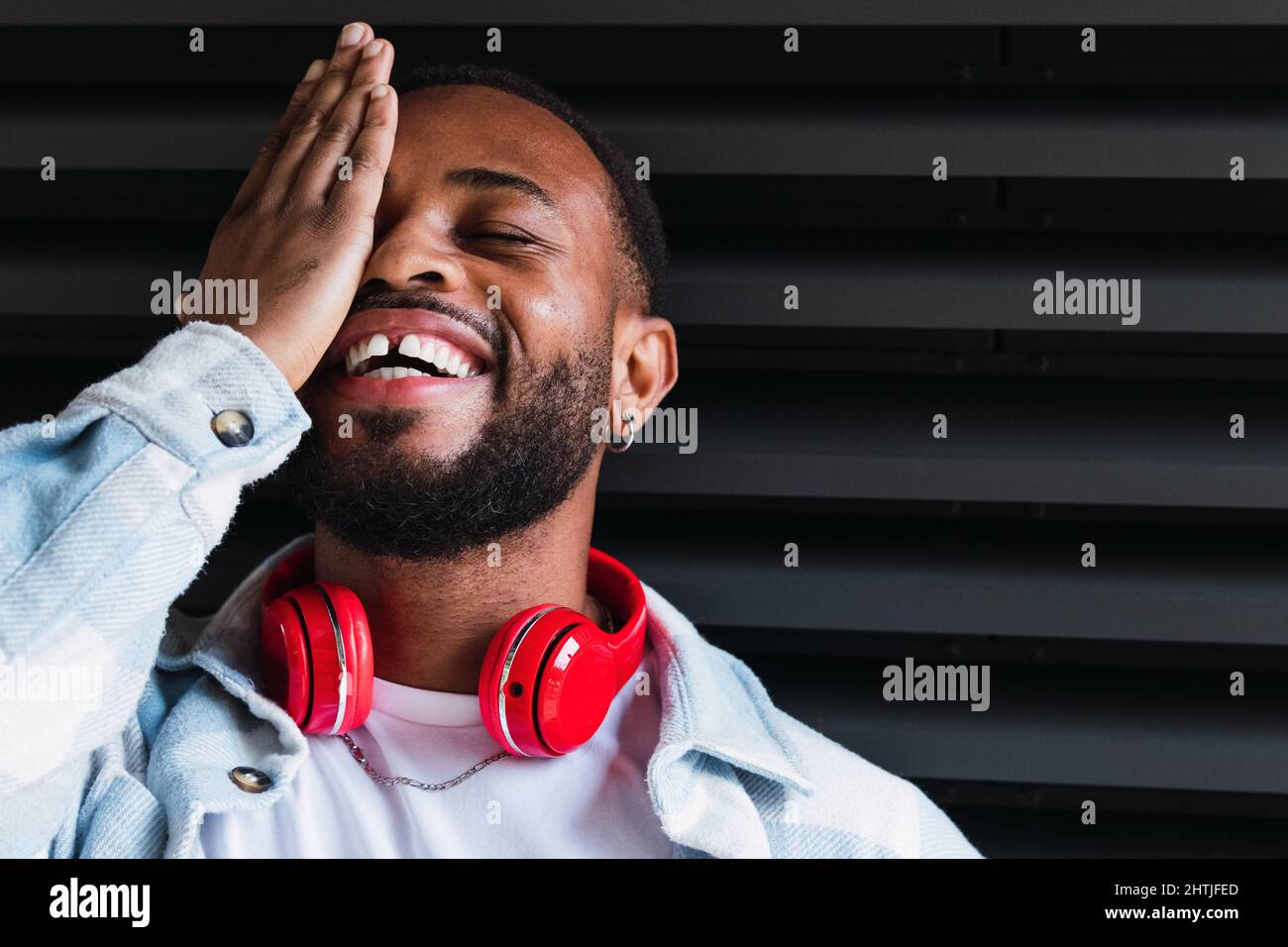 Homme afro-américain à barbe avec casque sans fil rouge moderne couvrant un œil fermé tout en restant debout sur fond noir dans la rue Banque D'Images