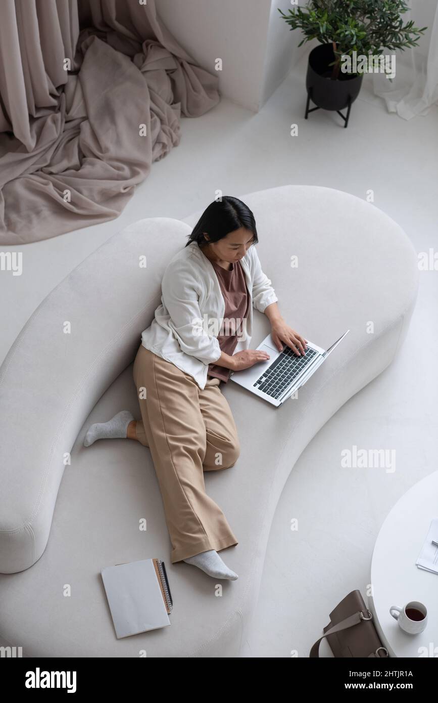 Jeune femme asiatique sérieuse dans des vêtements décontractés allongé sur un canapé tout en réseau ou faisant des achats en ligne à la maison Banque D'Images