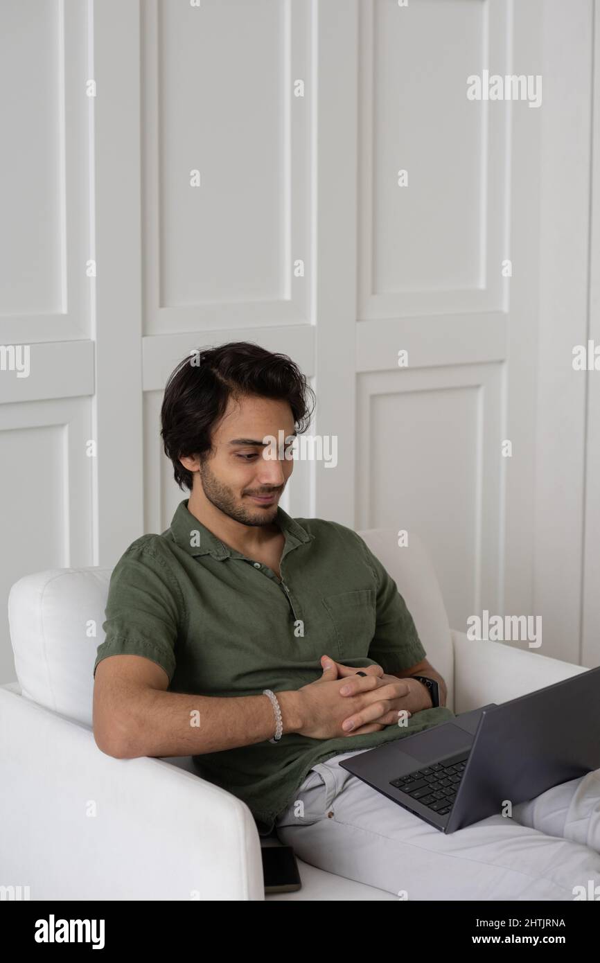 Jeune employé contemporain regardant l'écran d'un ordinateur portable sur ses genoux lors d'une réunion numérique tout en étant assis dans un fauteuil à la maison Banque D'Images