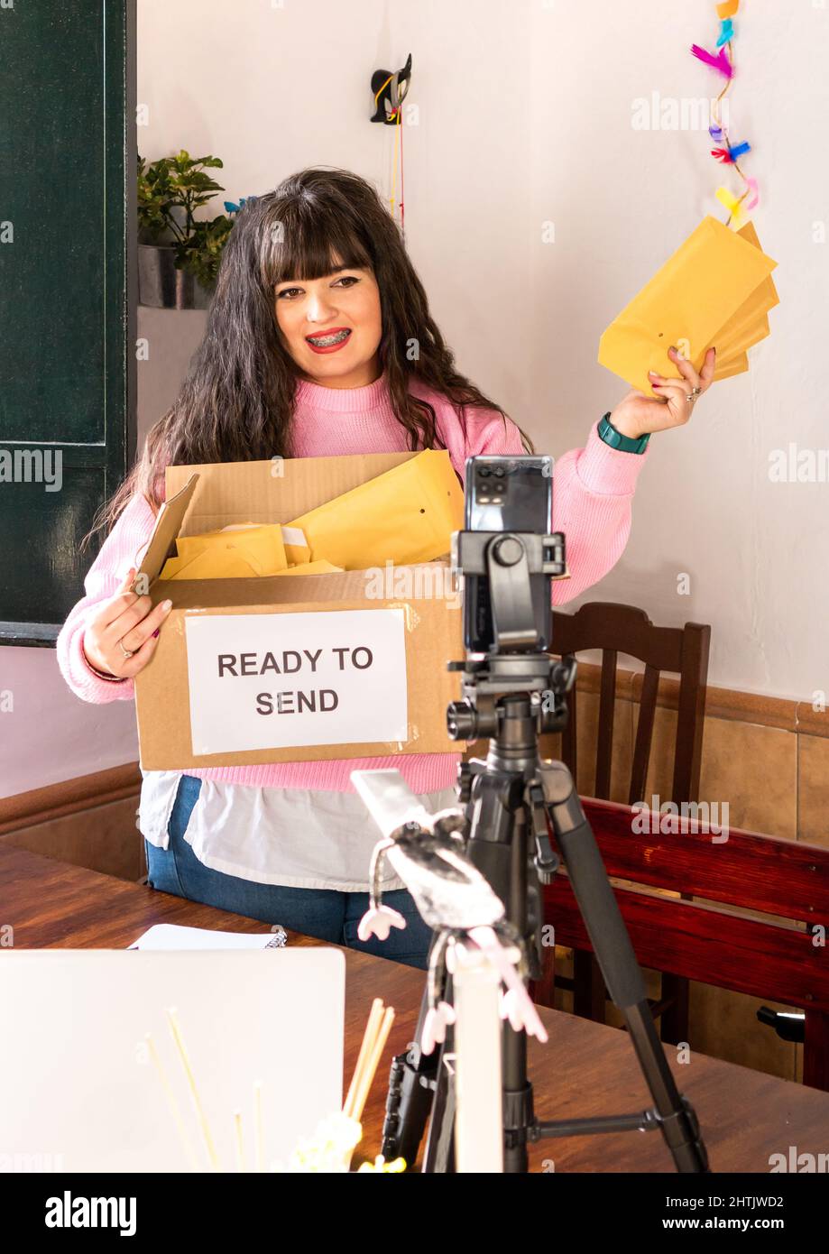 Femme blogger emballage enveloppes avec commandes dans carton pour livraison tout en enregistrant la vidéo sur téléphone portable attaché sur trépied dans l'atelier d'artisanat Banque D'Images