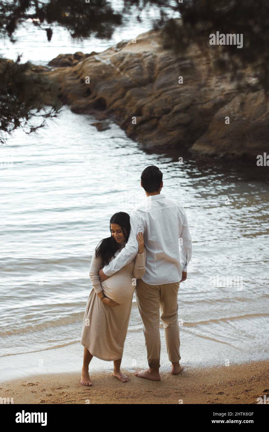 Jeunes femmes enceintes souriant et embrassant son mari sur la plage, le mari tient son ventre enceinte et regarde à l'horizon. Banque D'Images