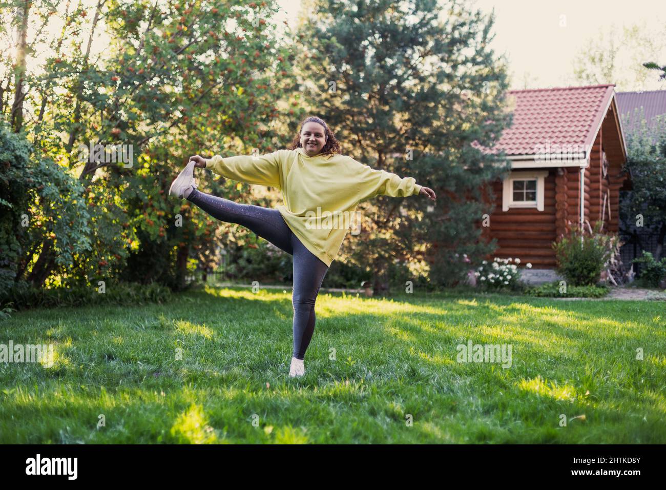 Femme adulte souriant sur l'herbe verte faisant des exercices dans les vêtements de sport avec un pied sur l'arrière-cour plein de lumière du soleil avec maison de campagne et de hauts arbres dedans Banque D'Images