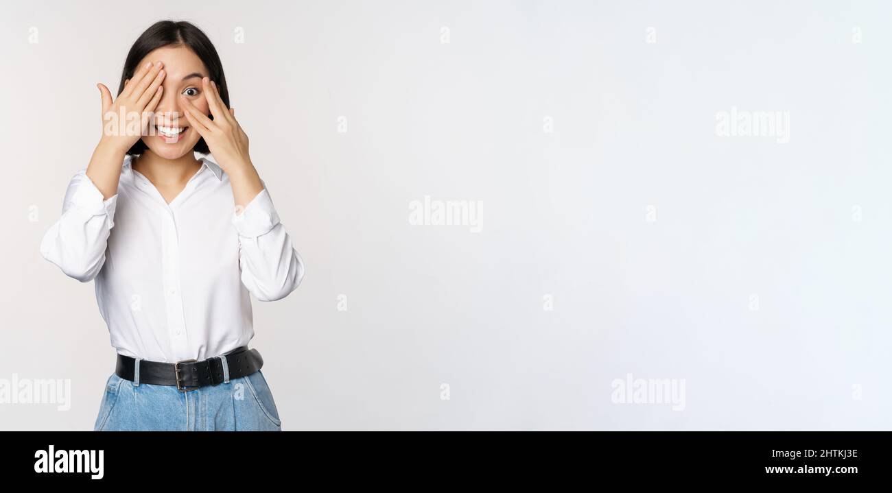 Image de la jeune femme asiatique ferme les yeux et sourit, attend la surprise, anticipe, peeking à travers les doigts, se dresse sur fond blanc Banque D'Images