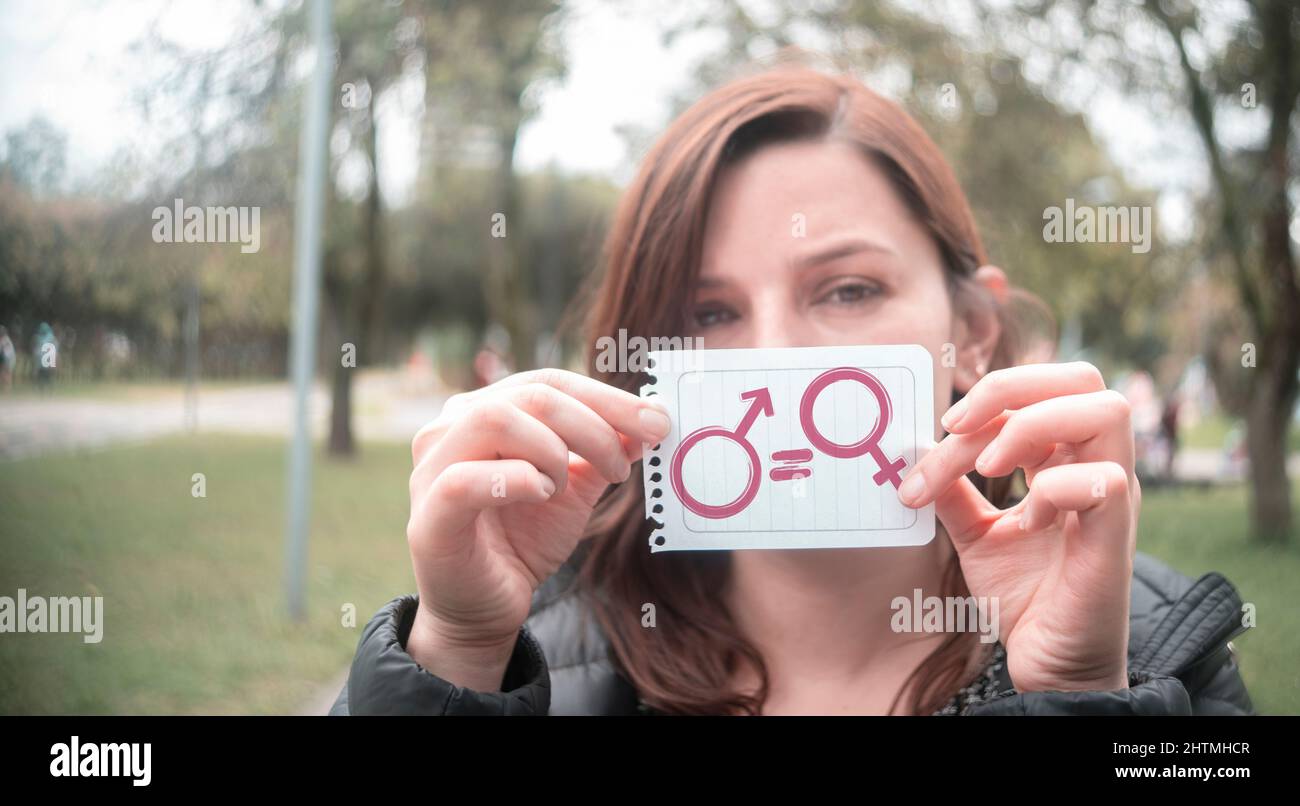 Belle femme hispanique au milieu d'un parc tenant dans la main un papier avec le message de l'égalité avec les icônes de l'homme et de la femme dans le front Banque D'Images