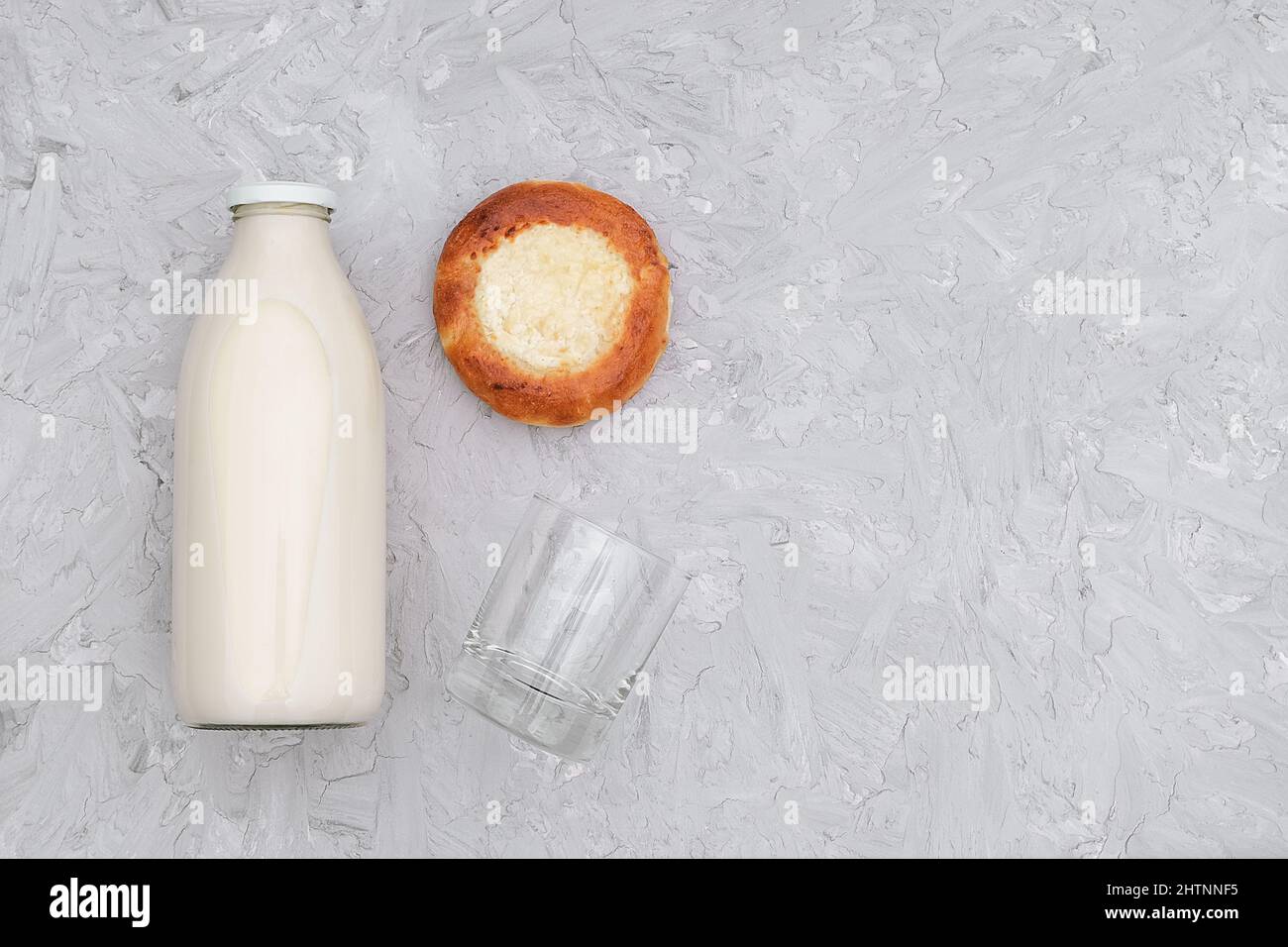 Lait dans une bouteille en verre, un verre vide et un petit pain fraîchement cuit sur fond gris avec espace de copie. Vue de dessus de la pose à plat. Banque D'Images