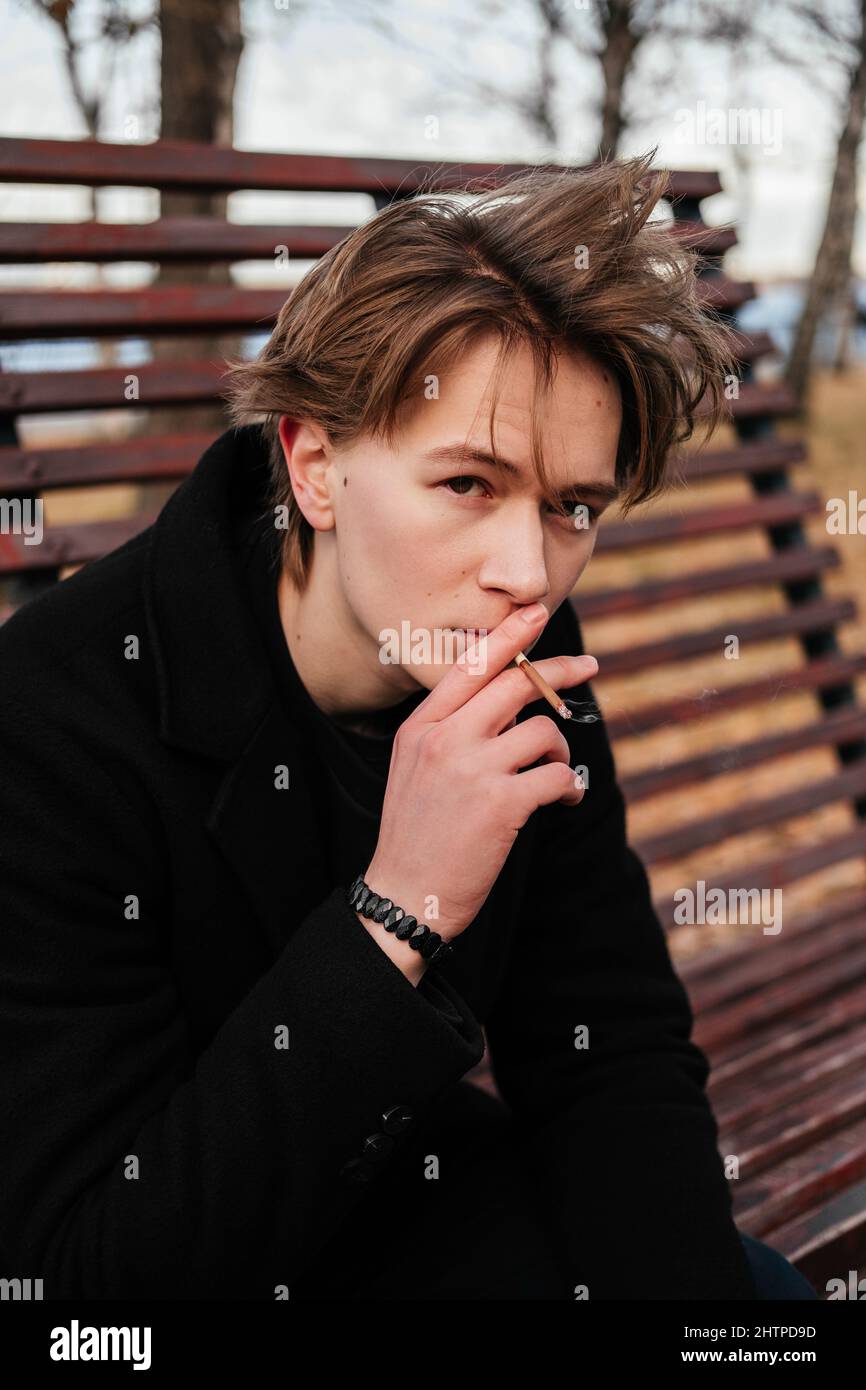 Jeune homme aux marques de naissance, moles, verrue sur le visage en manteau noir assis sur un banc en bois et fuyant une cigarette sur une rue de ville et regardant loin. Handso Banque D'Images