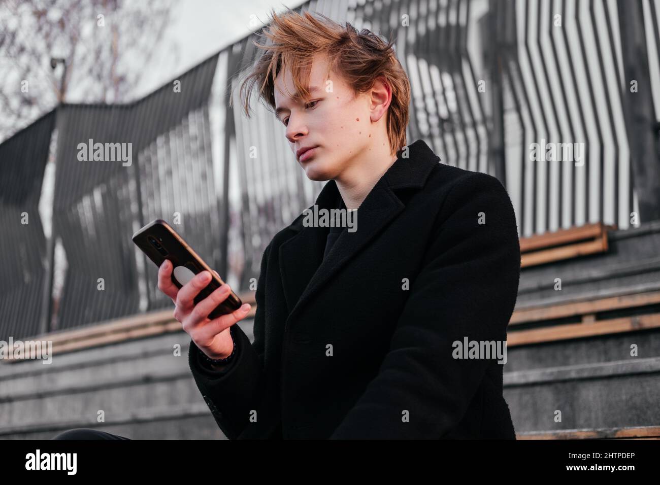 Jeune homme aux marques de naissance, moles, verrue sur le visage en manteau  noir avec smartphone dans les mains assis seul sur un banc en bois dans une  rue. Travailler sur le