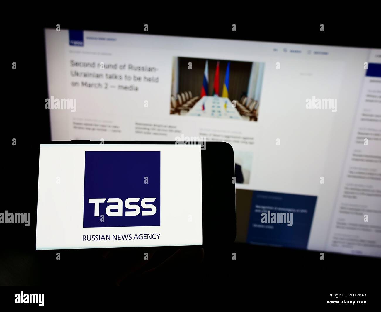 Personne tenant un téléphone portable avec le logo de l'agence de presse russe TASS (TACC) à l'écran en face de la page Web d'affaires. Mise au point sur l'affichage du téléphone. Banque D'Images