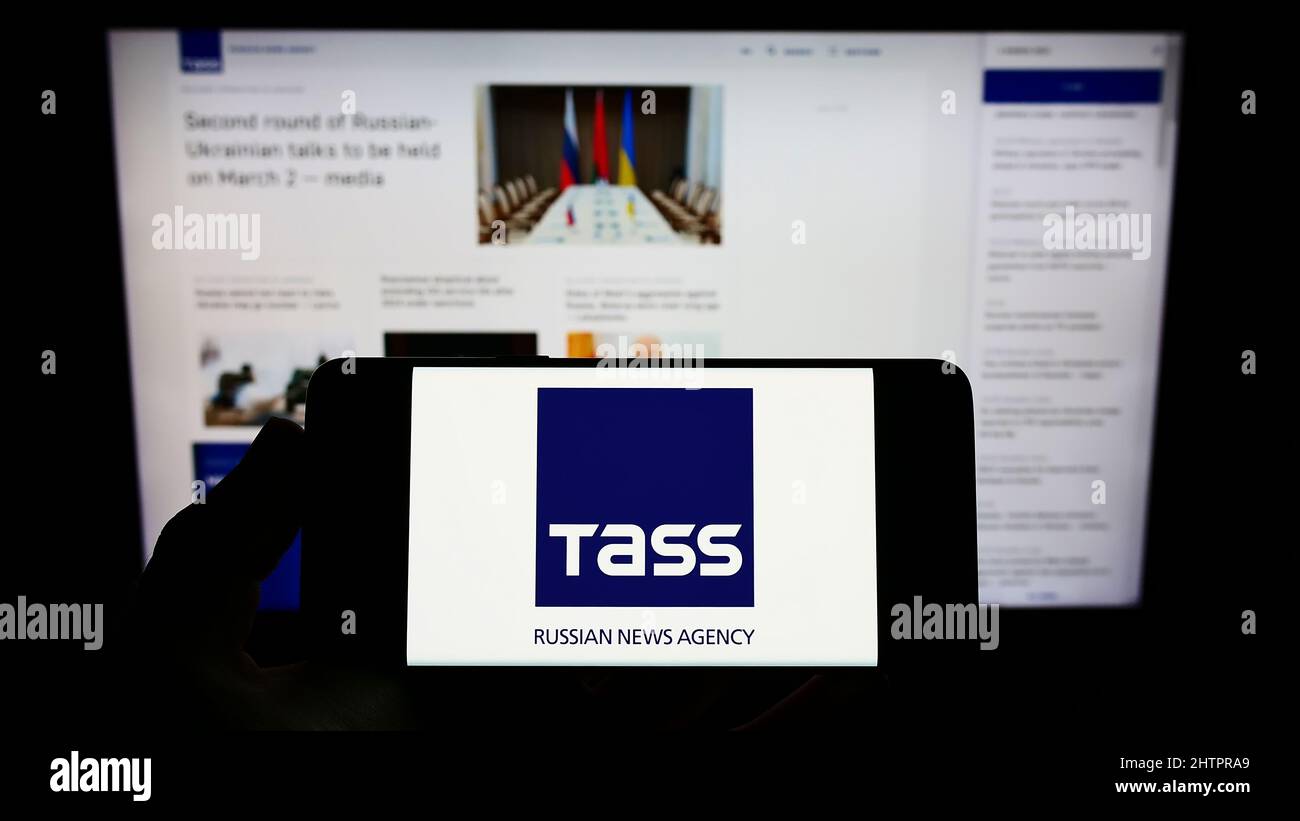 Personne tenant un téléphone portable avec le logo de l'agence de presse russe TASS (TACC) à l'écran en face de la page Web d'affaires. Mise au point sur l'affichage du téléphone. Banque D'Images