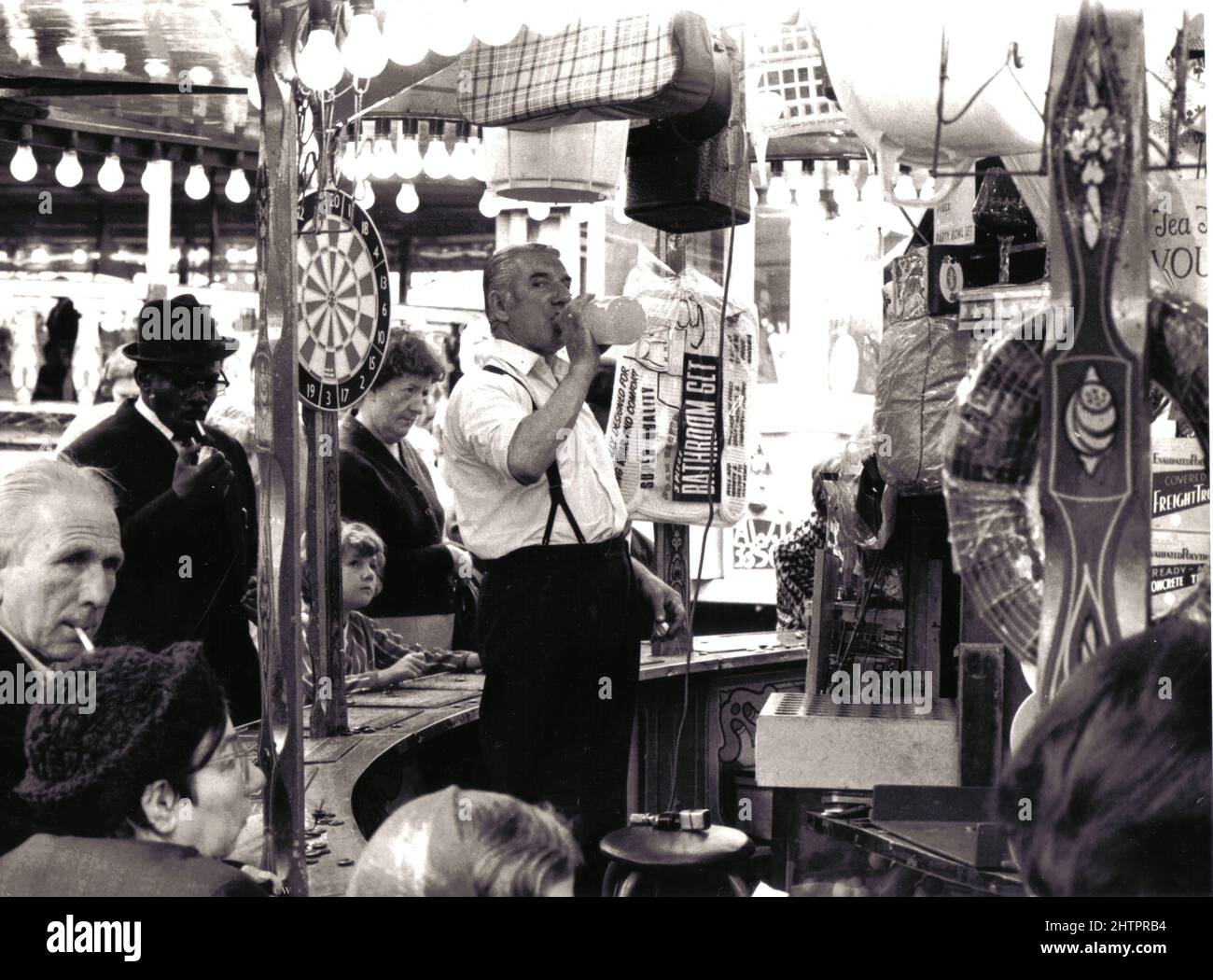 Un détenteur de stalle prend une boisson de bienvenue à son stand de bingo typique du parc des expositions à Manningham, Bradford, West Yorkshire, Angleterre, au début des années 1960. Divers prix tels qu'un ensemble de salle de bains et un dartboard peuvent être vu suspendu sur les supports de stalle circulaires. Banque D'Images