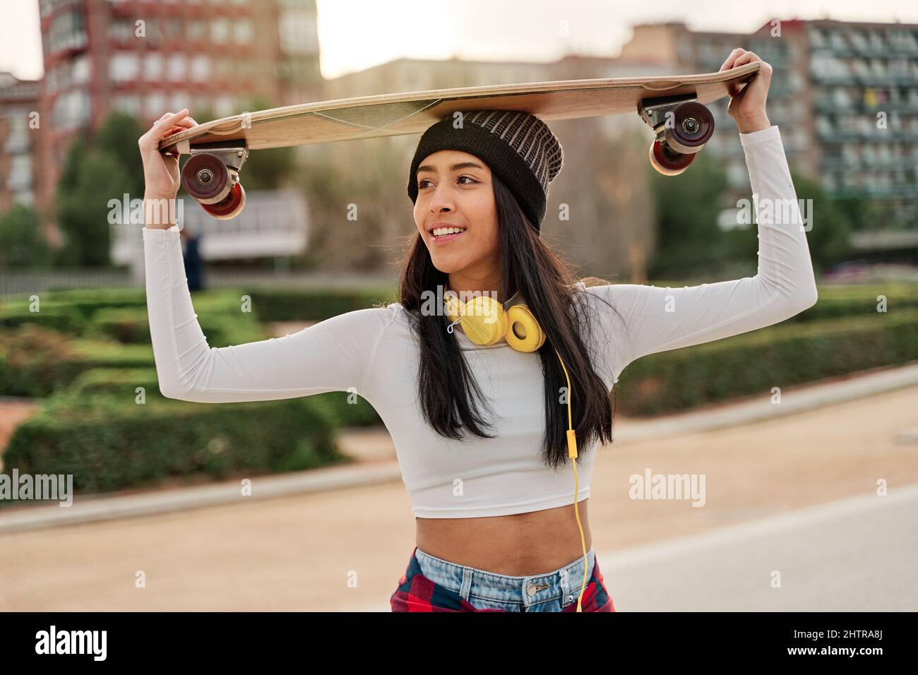 Latina femme dans des vêtements décontractés posant avec le skateboard sur sa tête Banque D'Images
