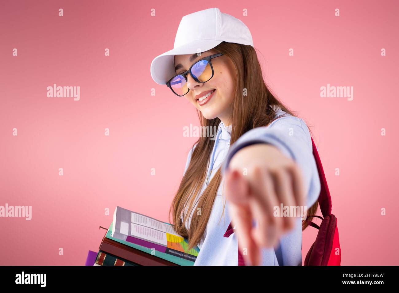 Cool jolie blonde fille d'école souriant portant des lunettes et une  casquette tenant des livres et pointant avec un doigt sur l'appareil photo  Photo Stock - Alamy