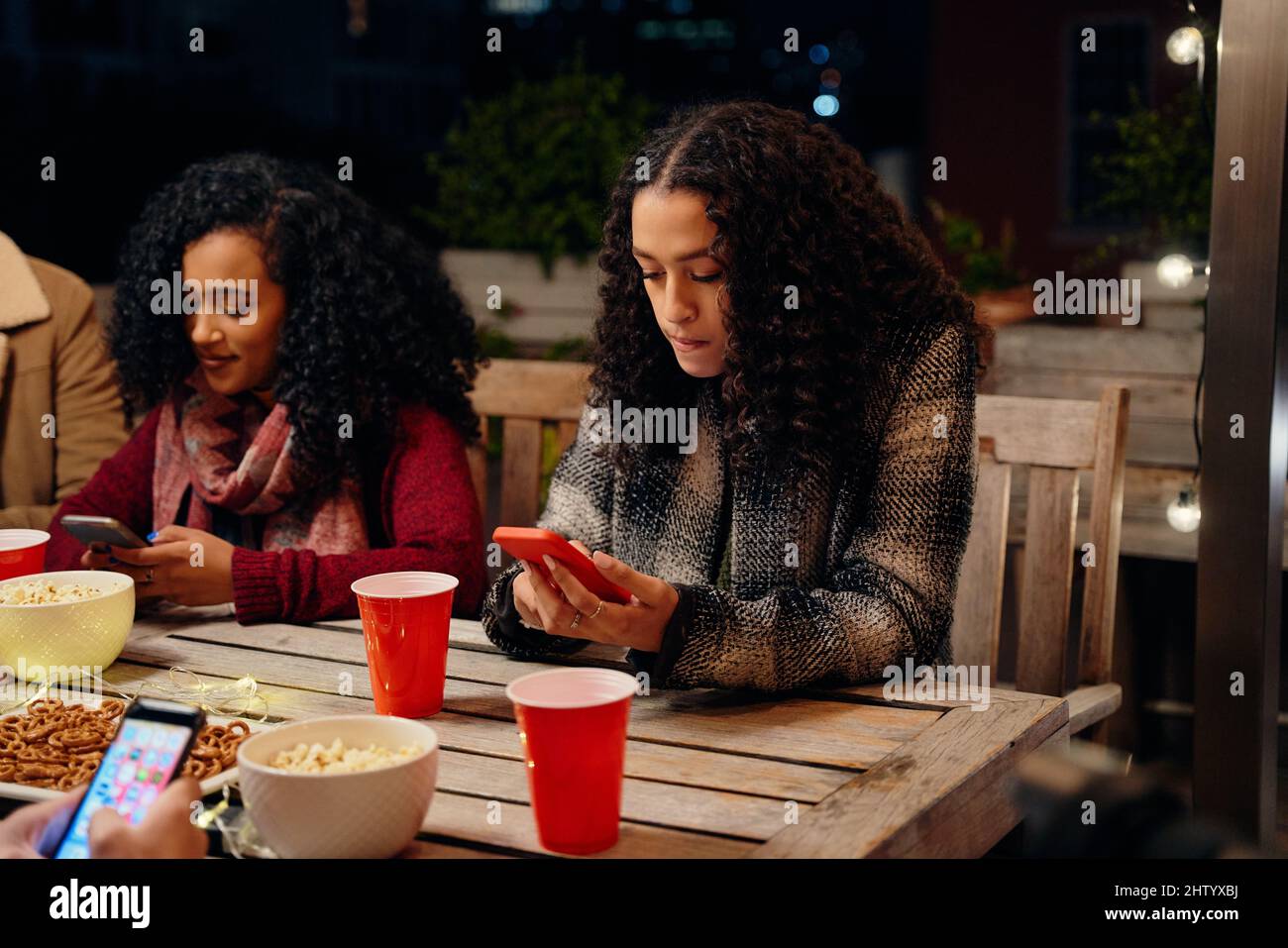 Deux jeunes femmes biraciales, assises à une fête, envoyant un SMS sur son téléphone. Anti-social, utilisant la technologie à la fête sur le toit Banque D'Images