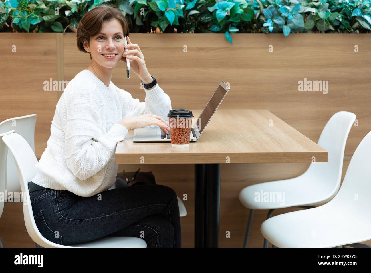 Jeune belle jolie brune femme portant un élégant sweat-shirt blanc assis dans un centre commercial à une table et travaillant à un ordinateur portable, en utilisant Banque D'Images