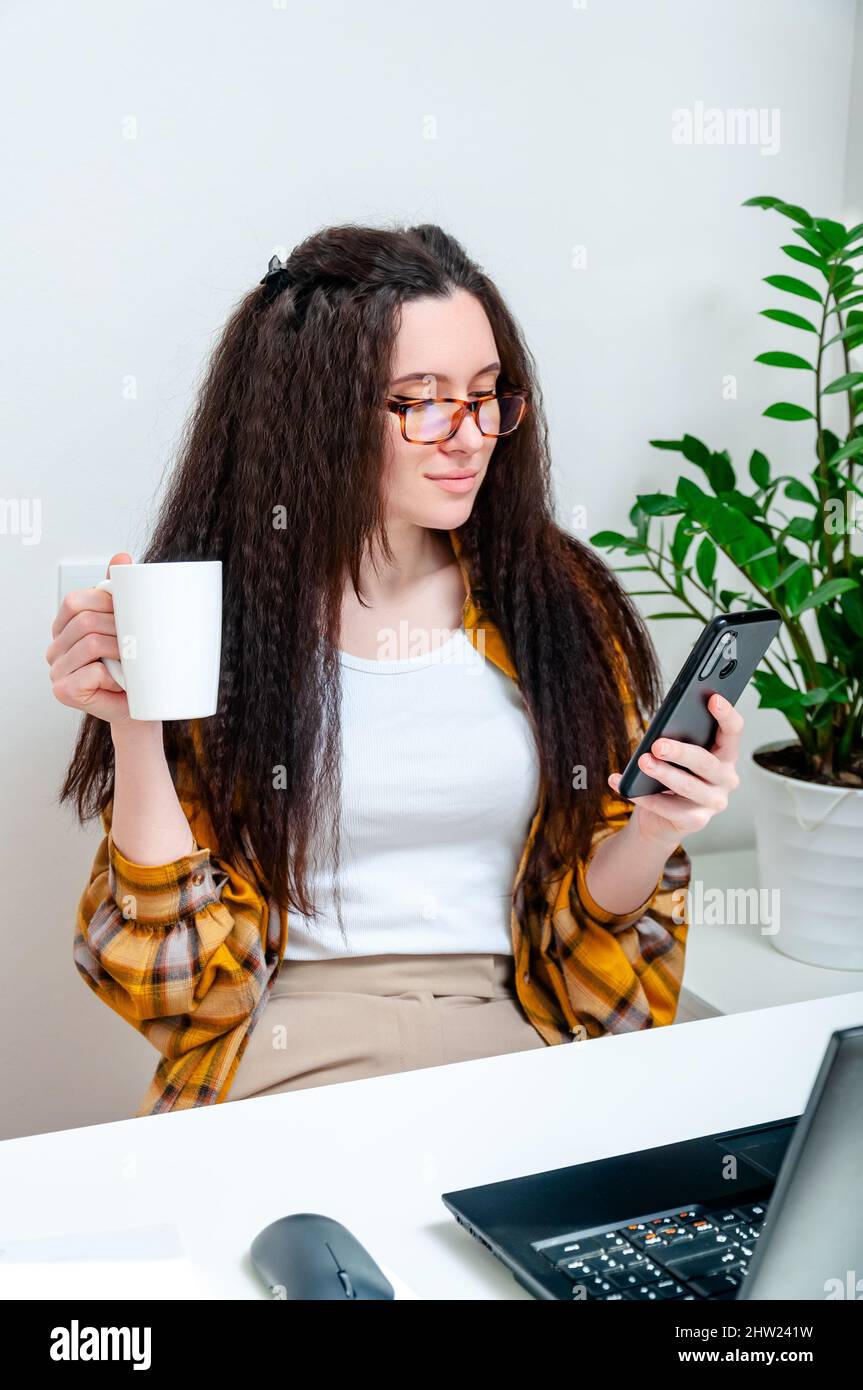 Une belle femme en verre boit du café pendant une pause et utilise un smartphone sur son lieu de travail. Femme travaillant à la maison, en cours de formation, regardant un webinaire, o Banque D'Images