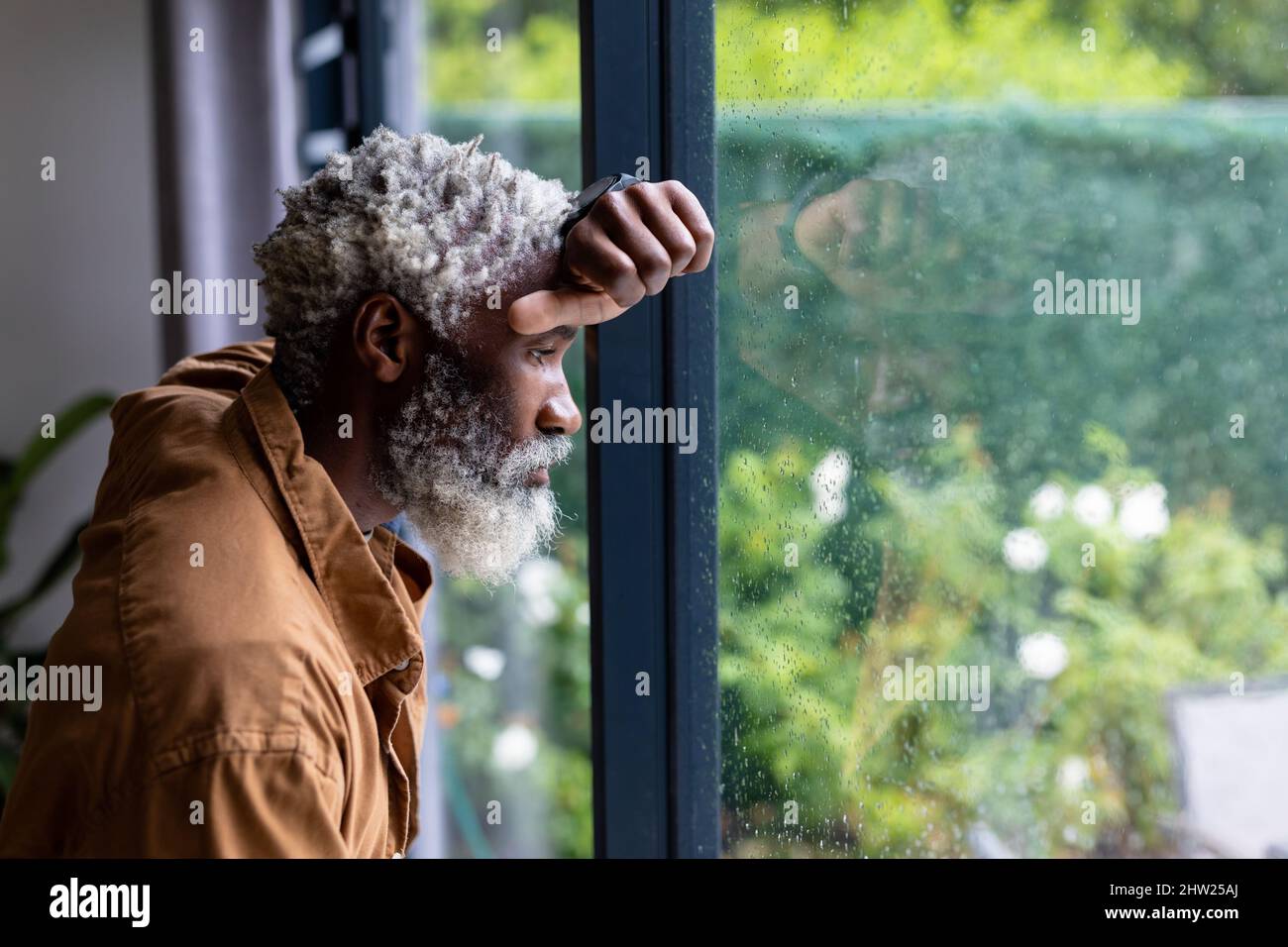 Ennuyé ancien africain américain à la retraite, pendu sur une fenêtre en verre tout en contemplant chez lui Banque D'Images
