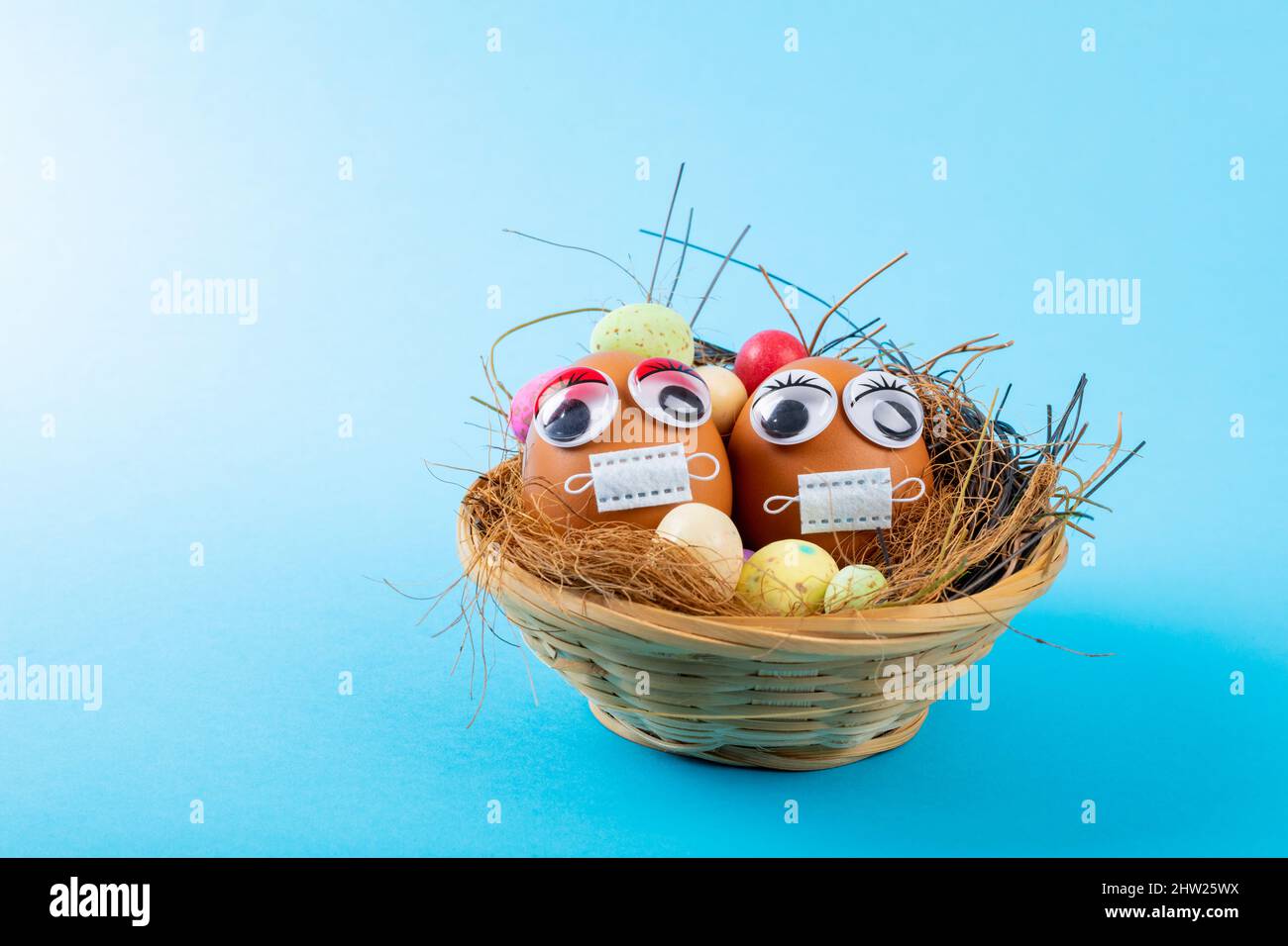 Gros plan des œufs de pâques avec des yeux en forme de poule et un masque avec des bonbons et des pailles dans le nid sur fond bleu Banque D'Images