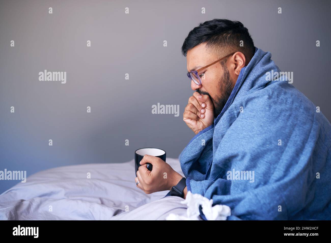 Un homme tousse dans un lit enveloppé dans une couverture contenant une boisson chaude. Copier l'espace Banque D'Images