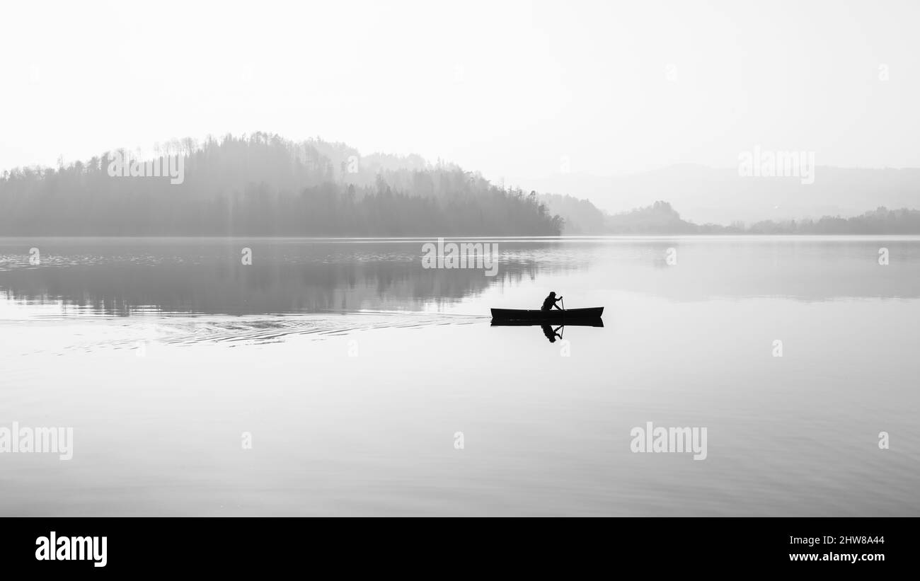 Brouillard sur le lac. Dans un miroir de réflexion d'eau calme. Homme avec une pagaie dans le bateau. Noir et blanc. Banque D'Images