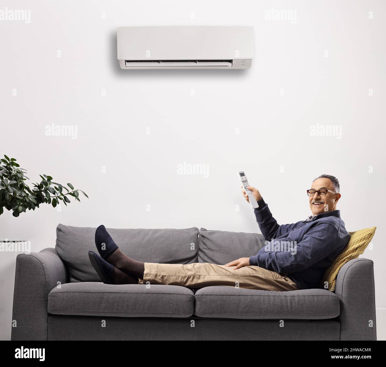 Homme mûr posé sur un canapé et mettant la climatisation avec une télécommande isolée sur fond blanc Banque D'Images