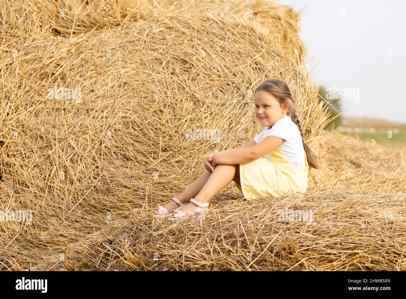 Portrait d'une petite fille heureuse avec les yeux calmés du soleil et long tresse sur la tête regardant loin assis sur des haystacks. Temps hors de la ville dans le pays Banque D'Images