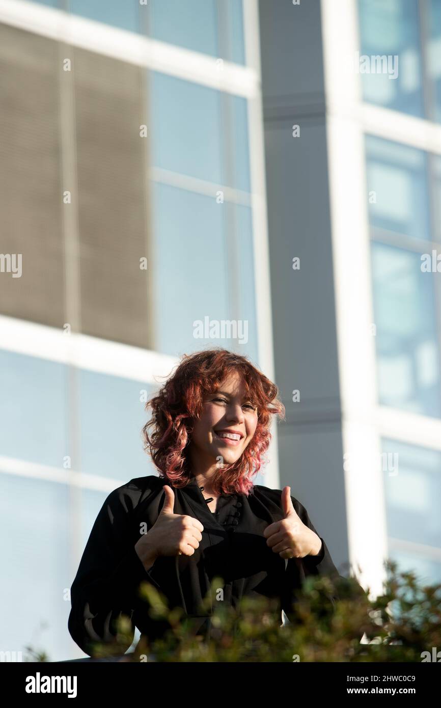 femme dans ses années 30 avec des cheveux rouges bouclés souriant à l'appareil photo avec les pouces vers le haut à l'extérieur Banque D'Images