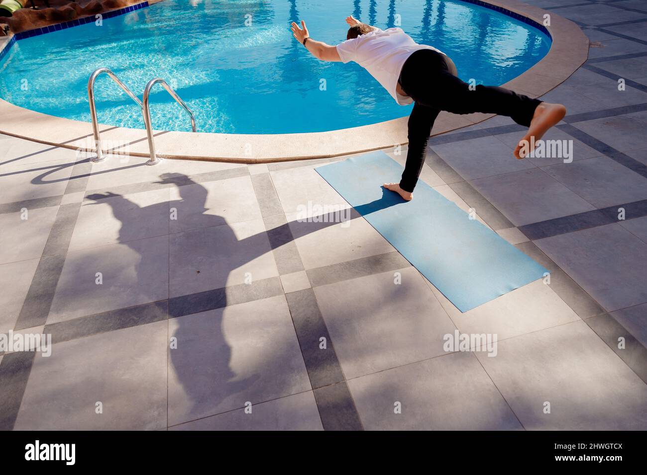 Asana debout sur une jambe, méditation yoga homme caucasien en chemise blanche sur fond piscine, palmiers et lumière du soleil. Banque D'Images