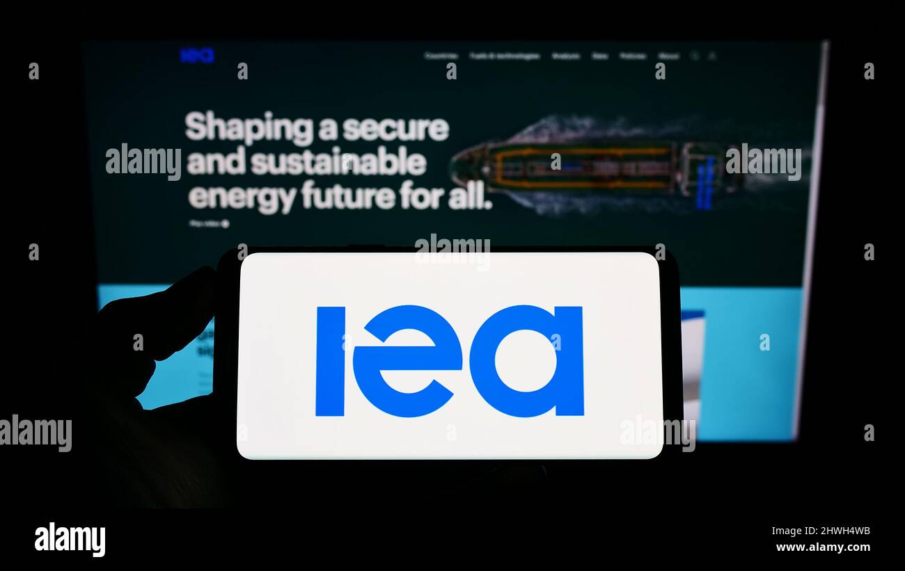 Personne tenant un téléphone portable avec le logo de l'organisation Agence internationale de l'énergie (AIE) sur l'écran en face de la page Web. Mise au point sur l'affichage du téléphone. Banque D'Images