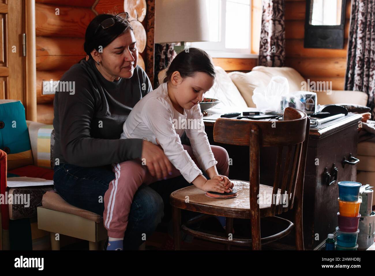 Jeune fille assise à genoux de la mère enseignant à la fille de poncer et enlever la peinture de vieux meubles avec papier de verre dans l'atelier à la maison. Durable Banque D'Images