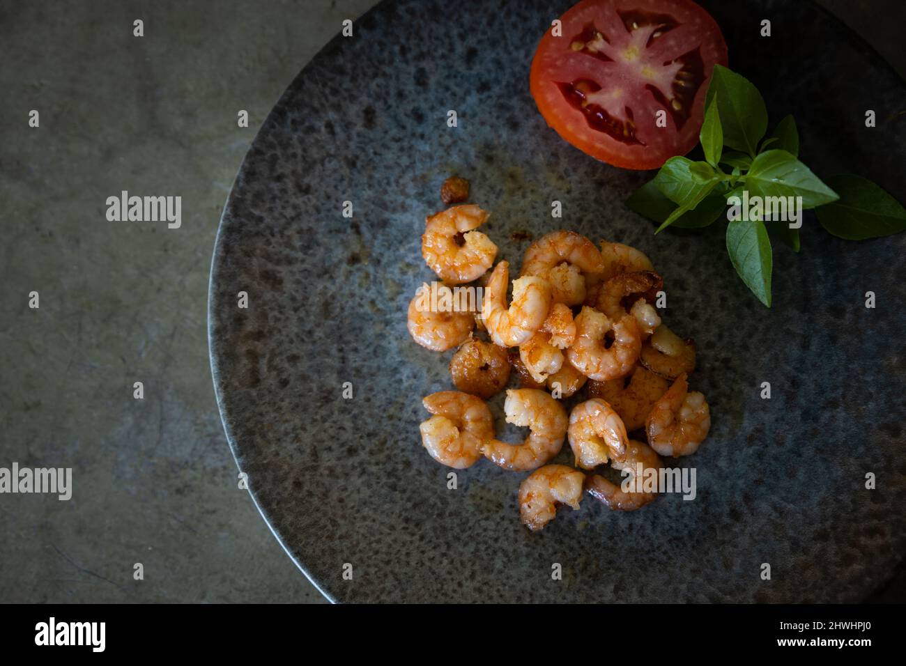 De délicieuses crevettes fraîchement cuites sur une assiette grise à côté d'une tomate et d'un basilic. Espace libre à écrire. Fond gris neutre. Banque D'Images