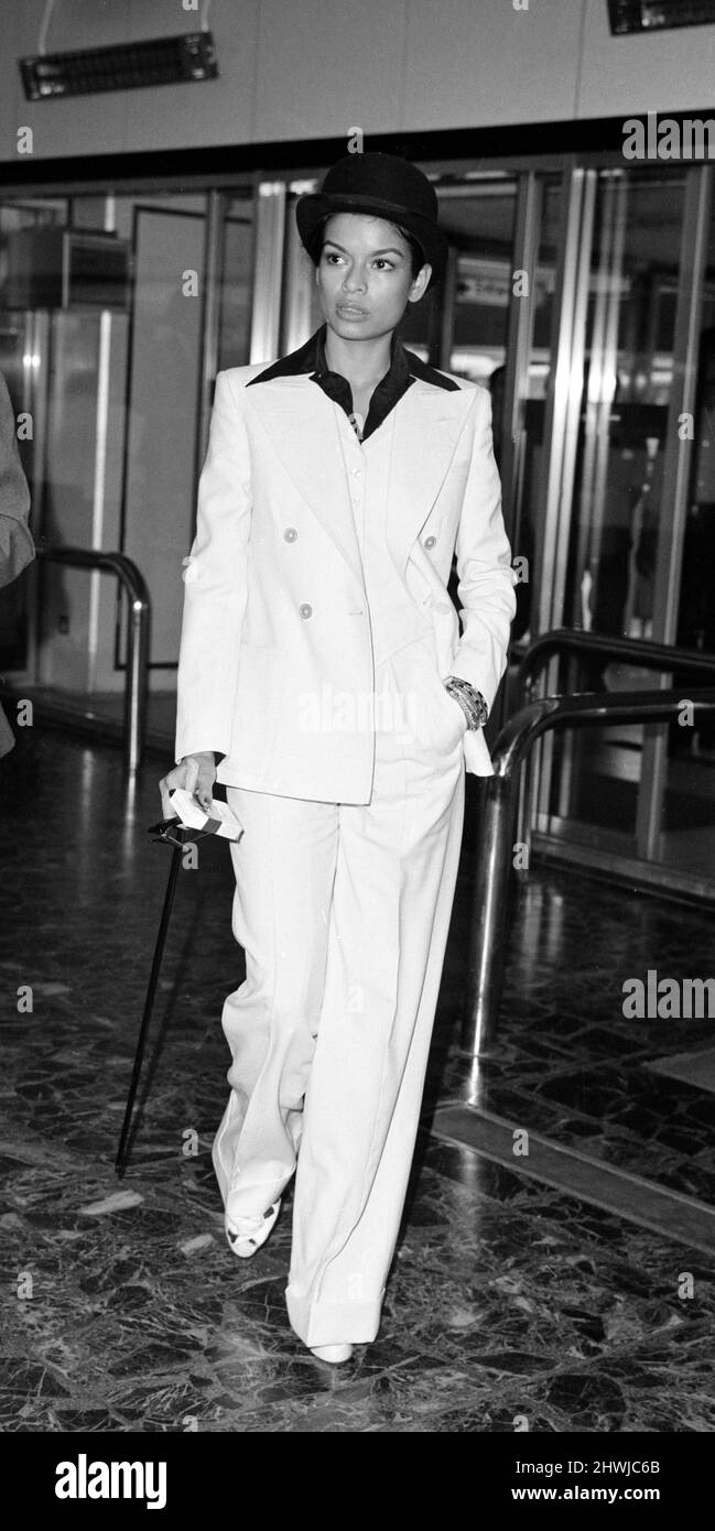 00092485 Bianca Jagger, épouse Mick Jagger, en costume blanc et chapeau de  lanceur avec canne à tête argentée alors qu'elle arrive à l'aéroport de  Heathrow à Londres pour un vol à New
