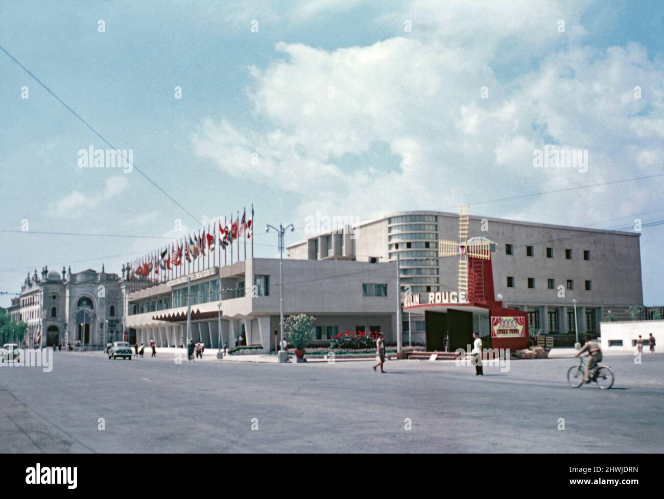 Une 1953 vue du Palazzo del Cinema sur le Lungomare Marconi, Lido di Venezia, Venise, Italie, le siège principal du Festival International du film de Venise – l'année 14th a eu lieu cette année-là. Un kiosque en forme de moulin à vent fait la promotion de ‘Poulin Rouge’, un film de 1952 réalisé par John Huston € il a gagné le Lion d'argent. Le Palazzo del Cinema a été construit dans le style moderniste, conçu par Luigi Quagliata, et ouvert en 1937. Il y a une salle et un cinéma de projection de 1032 places (à droite). Il a été agrandi en 1952 lorsque la façade de Quagliata a été construite (au centre à gauche) – une photographie vintage de 1950s. Banque D'Images Une 1953 vue du Palazzo del Cinema sur le Lungomare Marconi, Lido di Venezia, Venise, Italie, le siège principal du Festival International du film de Venise – l'année 14th a eu lieu cette année-là. Un kiosque en forme de moulin à vent fait la promotion de ‘Poulin Rouge’, un film de 1952 réalisé par John Huston € il a gagné le Lion d'argent. Le Palazzo del Cinema a été construit dans le style moderniste, conçu par Luigi Quagliata, et ouvert en 1937. Il y a une salle et un cinéma de projection de 1032 places (à droite). Il a été agrandi en 1952 lorsque la façade de Quagliata a été construite (au centre à gauche) – une photographie vintage de 1950s. Banque D'Images