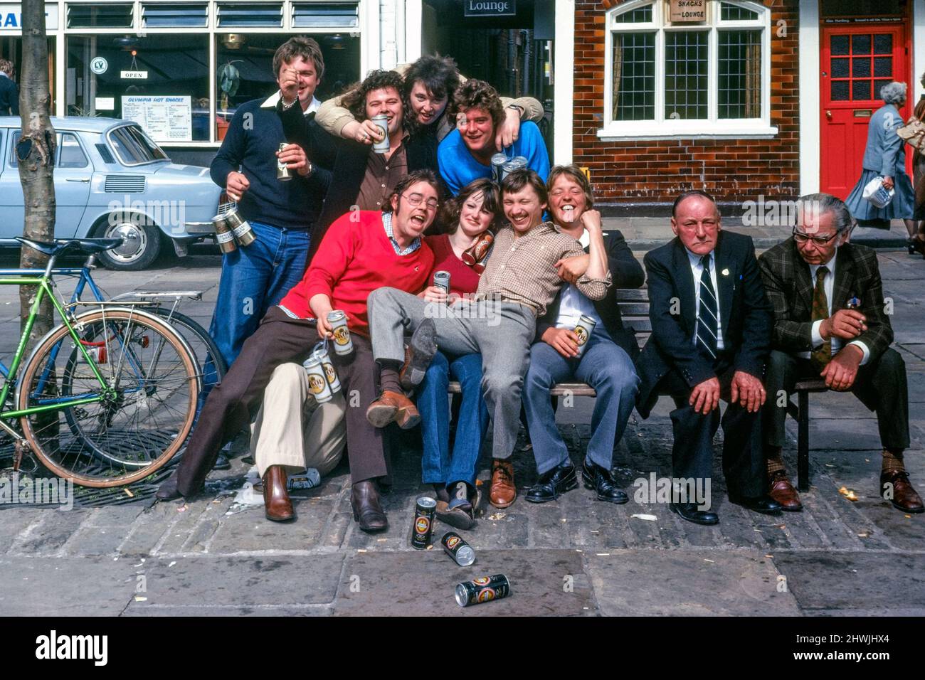 les jeunes hommes et les femmes solitaires transportant des canettes de bière se rassemblent sur le banc public à côté de deux hommes âgés tout en célébrant dans la rue 1980s york Banque D'Images