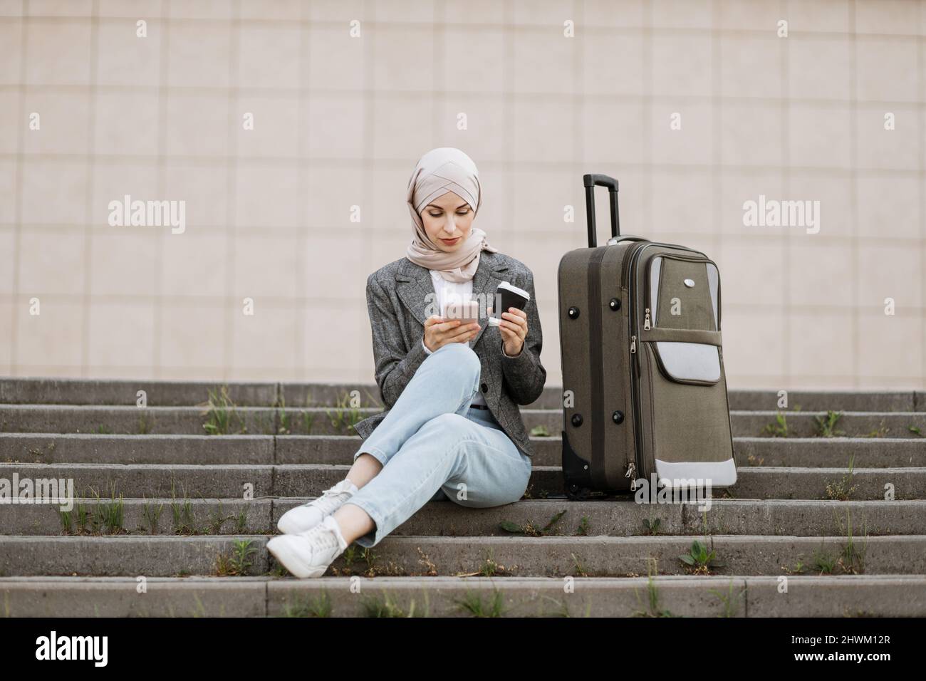 Jeune femme musulmane en hijab vêtue de jeans et de veste en utilisant le  téléphone et boire un café à emporter, tout en étant assise sur les  escaliers avec un sac de
