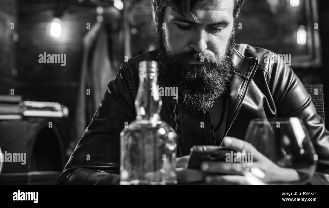 Style de vie de l'homme d'affaires moderne. Le beau barbu homme d'affaires boit cher whisky. Macho man défilement des nouvelles dans son smartphone. Banque D'Images