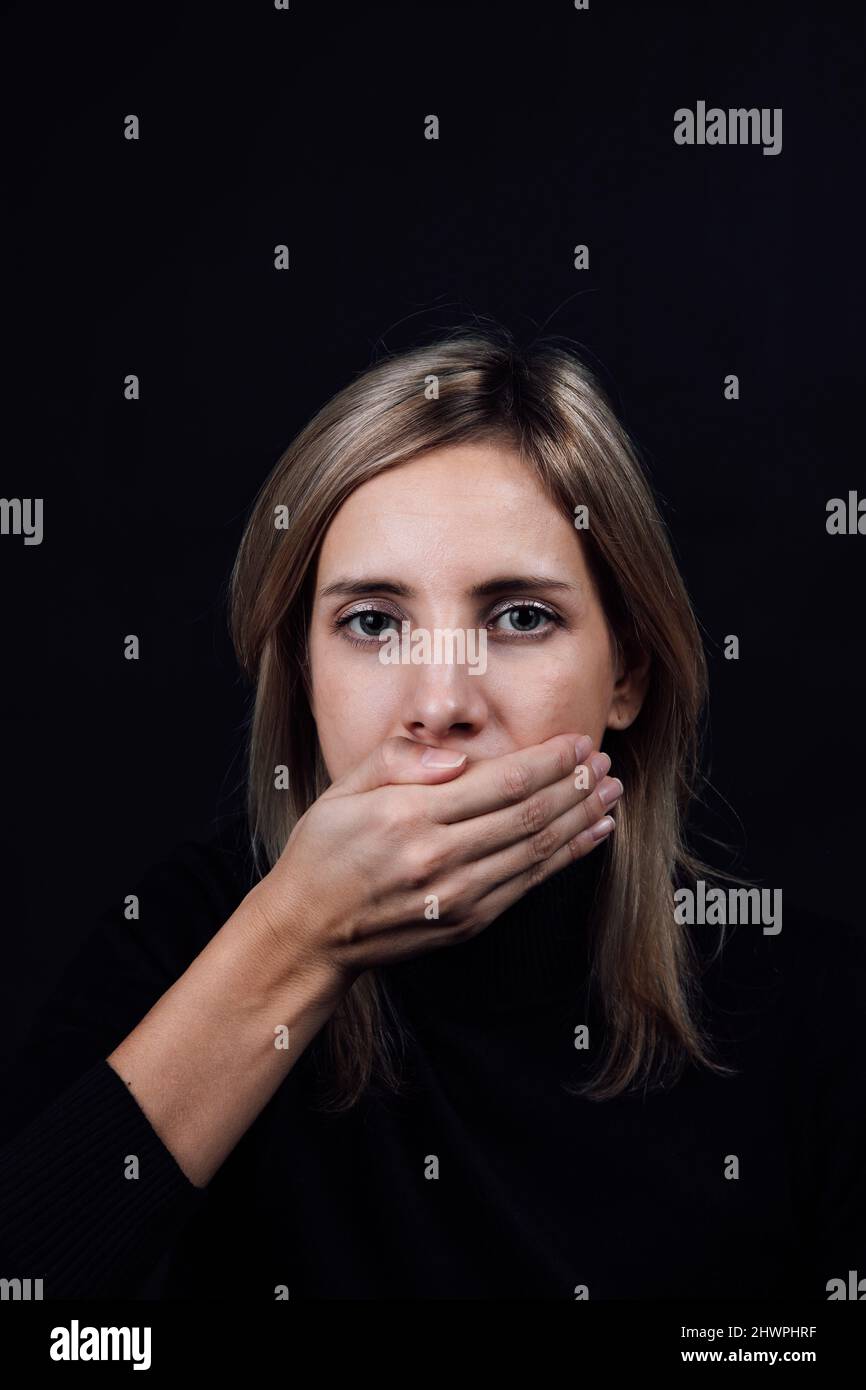 Femme blonde dépressive en blouse noire avec main couvrant la bouche et la rendant silencieuse sur fond noir. Victime de physique et psychologique Banque D'Images